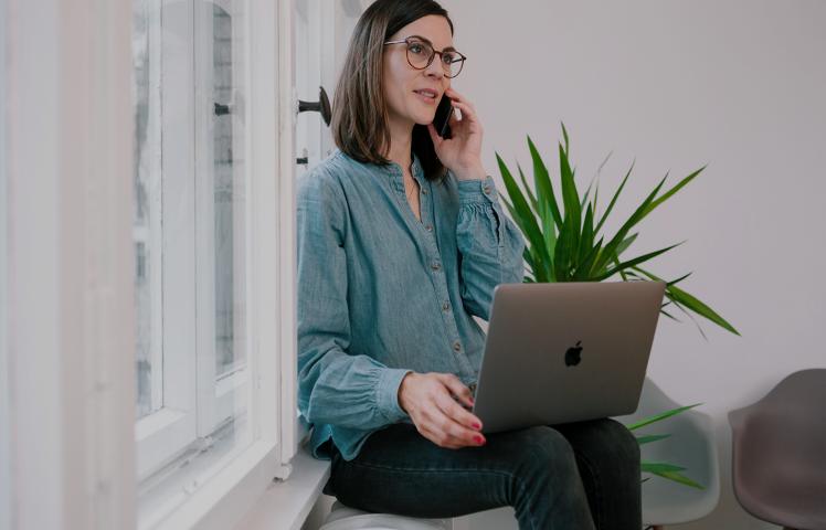 Eine Mitarbeiterin sitzt telefonierend auf der Fensterbank mit Laptop auf dem Schoß